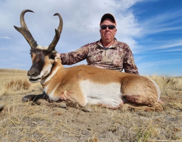 A hunter in camouflage attire posing beside his pronghorn antelope, showcasing its impressive horns in the golden grasses of Wyoming.